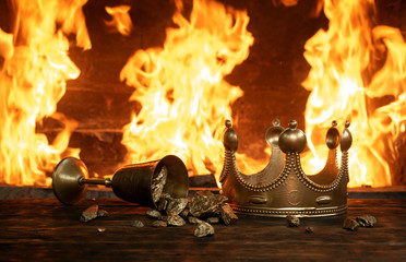 Golden crown and a goblet full of gold on the king table over burning fire background.