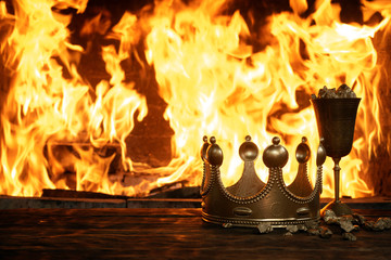Golden crown and a goblet full of gold on the king table over burning fire background.