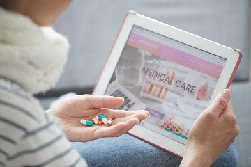 Close-up of unrecognizable woman holding pills in hand and using tablet while reading information about medications on internet
