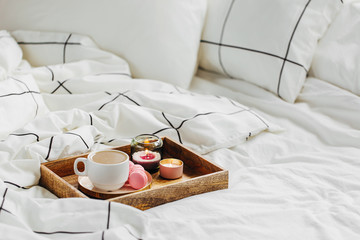 Wooden tray of coffee and candles on bed. White bedding sheets with striped blanket and pillow. Breakfast in bed. Hygge concept.