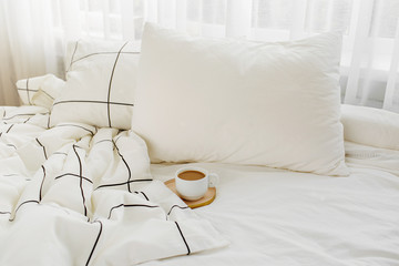 Cup of coffee on bed. White bedding sheets with striped blanket and pillow.  .