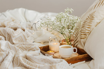 Fototapeta na wymiar Wooden tray of coffee and candles with flowers on bed. White bedding sheets with striped blanket and pillow. Breakfast in bed. Hygge concept.