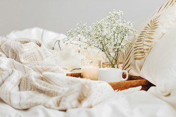 Wooden tray of coffee and candles with flowers on bed. White bedding sheets with striped blanket...