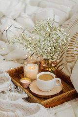 Wooden tray of coffee and candles with flowers on bed. White bedding sheets with striped blanket and pillow. Breakfast in bed. Hygge concept.