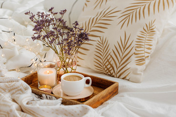 Wooden tray of coffee and candles with flowers on bed. White bedding sheets with striped blanket...