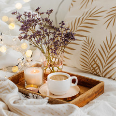 Wooden tray of coffee and candles with flowers on bed. White bedding sheets with striped blanket...