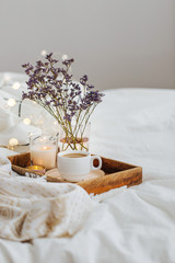 Wooden tray of coffee and candles with flowers on bed. White bedding sheets with striped blanket...