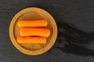 Group of three whole peeled orange baby cut carrot on bamboo plate flatlay on grey stone