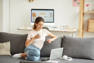 Positive attractive young woman sitting on comfortable sofa and drinking coffee while doing online...