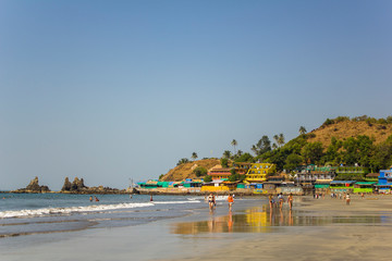 Fototapeta na wymiar Arambol, Goa/India - 04.01.2019: people walk along the sandy beach of the coast and swim in the ocean near the bright hotels near the hill with palm trees