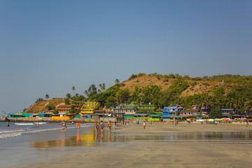 Fototapeta na wymiar Arambol, Goa/India - 04.01.2019: people walk along the sandy beach of the coast and swim in the ocean near the bright hotels near the hill