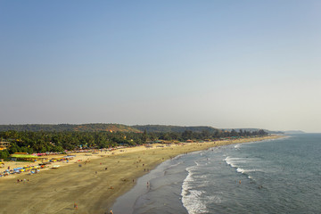 Arambol, Goa/India - 04.01.2019: people walk along the sandy beach of the sea coast and swim in the ocean against the backdrop of sun beds and palm jungle, aerial view
