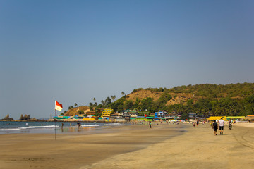 Fototapeta premium Arambol, Goa/India - 04.01.2019: people walk along the sandy beach of the seashore and bathe in the ocean against the background of bright multi-colored hotels near the hill