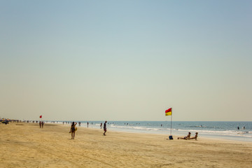 Obraz premium Arambol, Goa/India - 04.01.2019: sandy coast of the ocean coast with walking and bathing people and a lifeguard flag