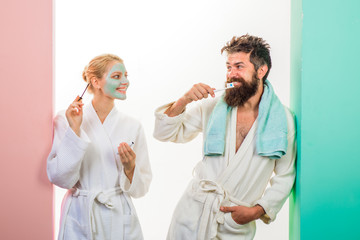 Obraz premium Morning preparation. Wife and husband preparing for work. Girl with cosmetic mask on face applying mascara. Couple. Woman applying mascara, man brushing teeth. Bearded man in bathrobe brushing teeth.