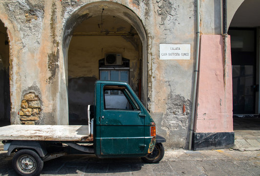 An old Ape car parked in front of the porches of Calata Cuneo with the street sign on a scraped wall, Oneglia, Imperia, Liguria, Italy