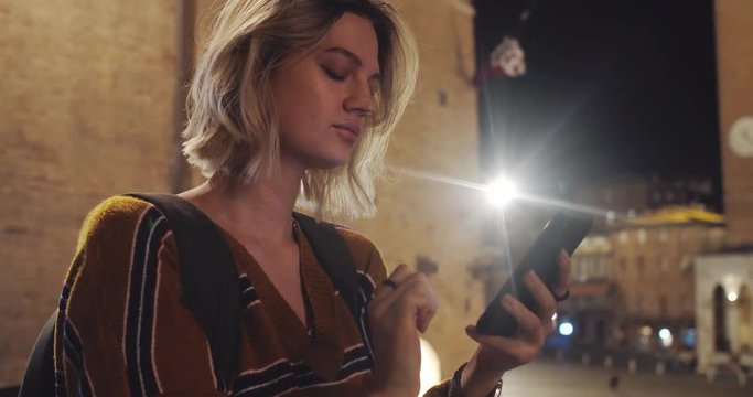 Portrait of sociable tourist girl using a mobile phone while exploring city at night. Close-up atractive pleasant girl reading on smartphone against gorgeous cityscape. Siena, Italy