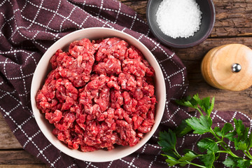 Fresh raw ground or minced beef meat in bowl, seasonings (salt and pepper) and fresh parsley on the side, photographed overhead (Selective Focus, Focus on the meat)