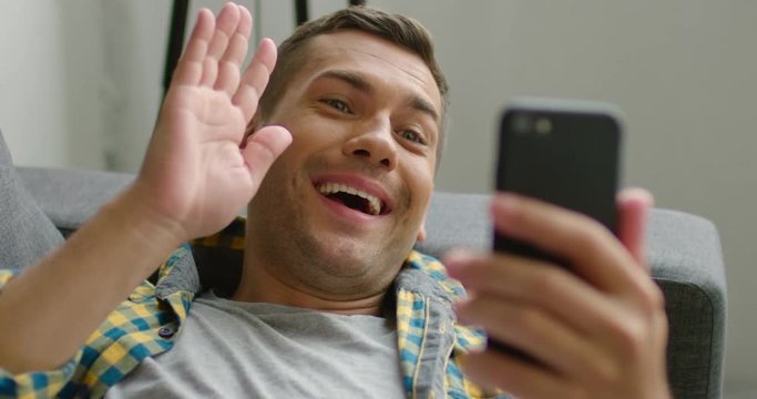 Very happy young man is talking via video application on his smartphone, smiling, waving with his hand, rasing his eyebrows, lying on a grey couch at home. Slow motion, 4K. Shot on RED camera.
