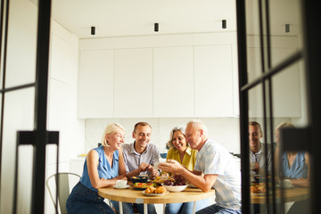 Group of cheerful senior people sitting around table in kitchen shot through open doors, copy space
