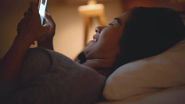 portrait of happy smiling young asian woman with smart phone while laying on bed in the evening