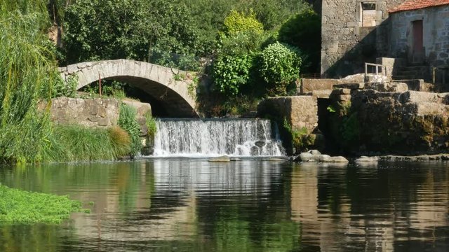 Ancient stone bridge over stream with waterfall and weeping willow tree. Beautiful summer scene on River Este in Portugal.