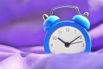Blue vintage alarm clock is on the bed wrapped in purple blanket. Arrows on dial show morning time in sunlight