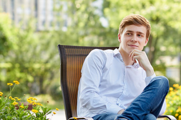 Young attractive man in white shirt sitting on brown chair in the park on green background - 279737800
