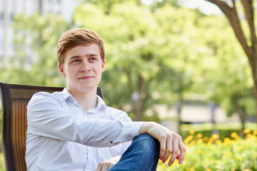 Young attractive man in white shirt sitting on brown chair in the park on green background - 279737852