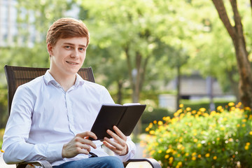 Young attractive man in white shirt sitting on brown chair in the park on green background - 279738026