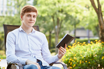 Young attractive man in white shirt sitting on brown chair in the park on green background - 279738267