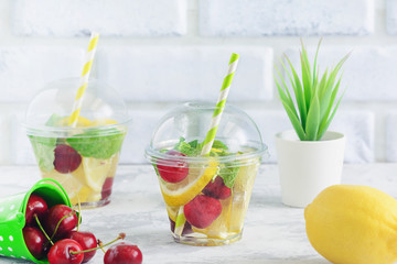 Water Glass with Organic Citrus Fruit Berry Mint. Healthy Party Beverage. Diet and Detox Concept. Plastic Cup of Cool Soda Lemonade. Ripe Sweet Cherry and Lemon on Light Kitchen Table