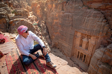 Asian man traveler sitting on carpet viewpoint in Petra ancient city looking at the Treasury or...
