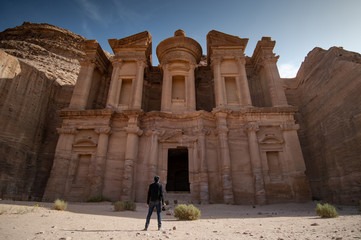 Young Asian man traveler and photographer holding camera standing at Ad Deir or El Deir, the monument carved out of rock in the ancient city of Petra, Jordan. Travel Middle East concept