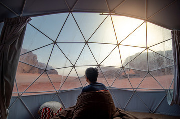 Tourist man staying in blanket in dome tent looking outside at Wadi Rum desert, famous natural attraction in Jordan. Travel Middle east concept