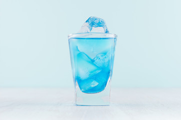 Blue curacao cocktail with ice cubes in luxury shot glass on white wood board and pastel mint color wall.