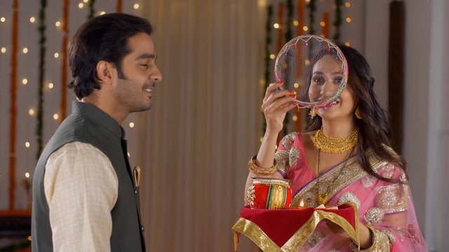 Young Indian couple happily performing Karwa Chauth traditions in traditional wear. Beautiful wife looking at the moon through a sieve and then to her good looking husband  following Karwa Chauth r...