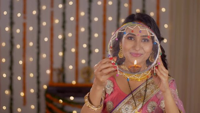 Pretty Indian woman happily looking into the camera through a sieve - Karwa Chauth concept. Beautiful married lady looking through a sieve at the moon and then at the camera  posing on the occasion...