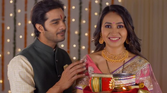 Indian husband-wife posing and smiling while looking into the camera - celebrating Karwa Chauth festival. Newly married couple celebrating their first Karwa Chauth festival - Dressed in traditional...