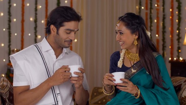 South Indian couple dressed in a traditional dress and drinking a hot tea together - Lifestyle. Loving couple spending time together at a festival at home. Interior decorated with lights and flower...