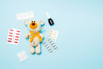 Children's knitted toy teddy bear with pills takes the temperature of a mercury glass thermometer on blue background. Child' disease concept. Top view, flat lay and copy space.