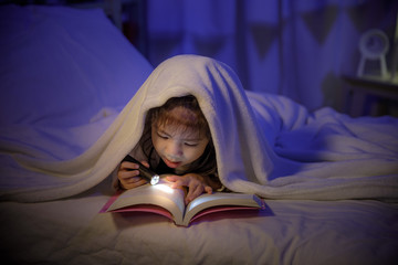 Child asian girl reading a book with flashlight under the blanket on bed in a dark bedroom at night before sleeping, Comfortable children at home concept