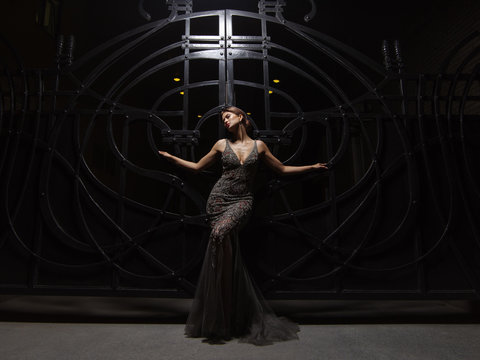 Passionate and attractive smartly dressed young woman in an expressive sparkling evening dress is posing near the big black wrought iron gate at night. Smart well-dressed glamour lady 