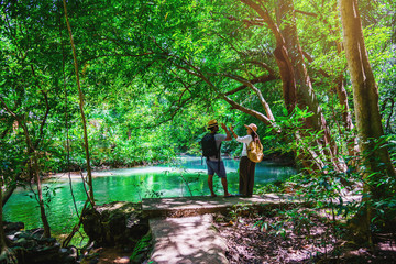 Naklejka na ściany i meble Travel couples with natural travel backpacks during the holidays. Couples traveling, relaxing in greens jungle and enjoying the beautiful Emerald pond. Tourism hiking nature study.
