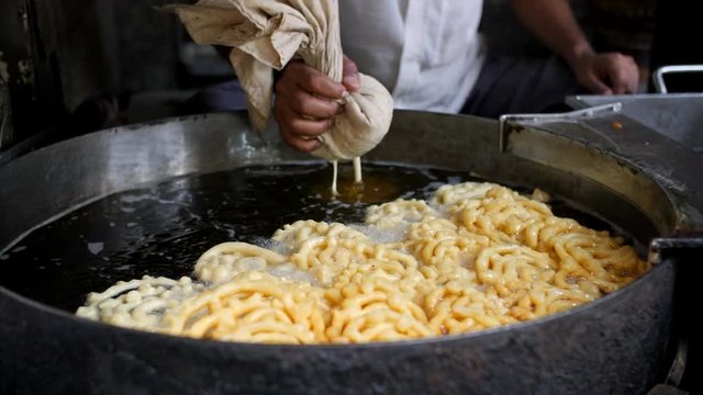An Indian sweet shop owner (Halwai) preparing and frying spiral jalebis (Indian Sweet Dish) in a huge oil container. 