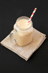 Mason glass jar mug filled with peanut butter banana smoothie on a black surface, low angle view. Close-up.