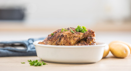 Whole chicken roasted in white dish with raw potatoes on kitched table. Home interior in the background