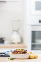 Whole chicken roasted in white dish with raw potatoes on kitched table. Home interior in the background
