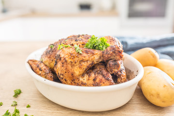 Whole chicken roasted in white dish with raw potatoes on kitched table. Home interior in the background
