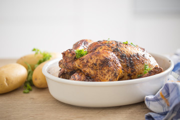Whole chicken roasted in white dish with raw potatoes on kitched table. Home interior in the background